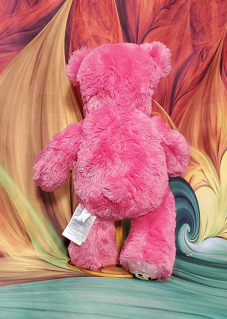 K&ouml;nnte beinhalten: Ein rosa Teddyb&auml;r mit weicher, pelziger Textur. Der B&auml;r wird von hinten betrachtet und zeigt seine Ohren, Arme und Beine. Ein Etikett ist am R&uuml;cken des B&auml;ren befestigt. Der Fu&szlig; des B&auml;ren hat ein kleines Design.