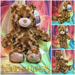 May include: A brown teddy bear with a curly fur texture, blue eyes, and a tan snout. The bear has a tag that says "The Lord of the Rings". The bear's paws have a map design and the words "The Lord of the Rings".
