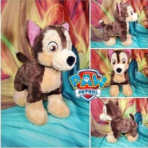 May include: Brown and tan plush toy dog with pink ears. The dog is wearing a blue collar with the words "PAW PATROL" on it. The dog is sitting on a blue and green surface.