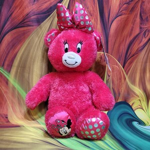 Puede incluir: Un oso de peluche rosa brillante con un gran lazo con lunares plateados. El oso tiene una cara sonriente, pesta&ntilde;as y Minnie Mouse en un pie. El otro pie tiene lunares turquesa.