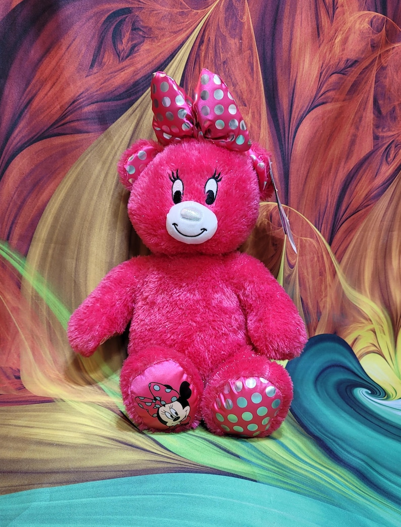 Puede incluir: Un oso de peluche rosa brillante con un gran lazo con lunares plateados. El oso tiene una cara sonriente, pesta&ntilde;as y Minnie Mouse en un pie. El otro pie tiene lunares turquesa.