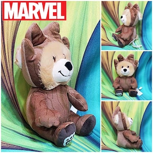 May include: A plush brown bear toy with a tree-like design, featuring a light brown face, ears, and chest. The bear has a black nose and a smiling mouth. The word "MARVEL" is in white block letters in the upper left corner.