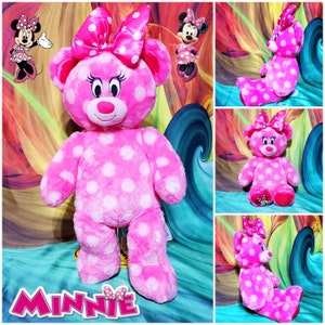 Op de afbeelding: Een roze pluche beer met witte stippen, met een grote roze strik. De beer heeft een roze neus, wimpers en een Minnie Mouse-ontwerp op zijn voet. Het woord "Minnie" wordt in roze letters weergegeven.