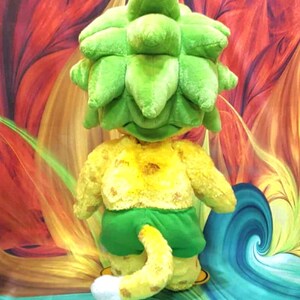 May include: A plush toy in the shape of a whimsical creature with a large, green, leafy head, yellow torso, green shorts, and a yellow tail with a white tip. The background features a colorful, abstract design with swirls of red, orange, and blue.