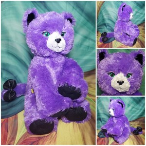 May include: A purple plush cat with black eyes and a white nose. The cat is sitting with its legs crossed and has a black bow on its tail.