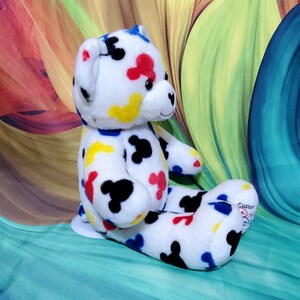 Pu&ograve; includere: Un orsacchiotto di peluche bianco con macchie colorate a forma di Mickey Mouse in rosso, giallo, blu e nero. L'orso &egrave; seduto con le gambe distese e ha un piccolo naso a forma di cuore.