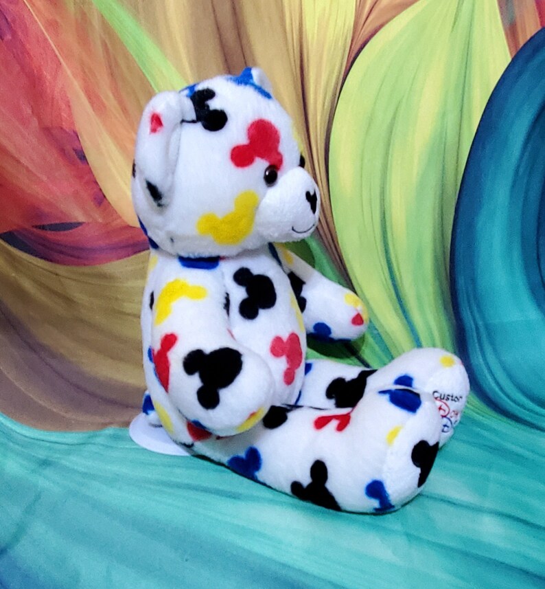 Pu&ograve; includere: Un orsacchiotto di peluche bianco con macchie colorate a forma di Mickey Mouse in rosso, giallo, blu e nero. L'orso &egrave; seduto con le gambe distese e ha un piccolo naso a forma di cuore.