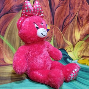 Puede incluir: Un oso de peluche rosa brillante con un gran lazo con lunares blancos. El oso tiene un hocico blanco, una cara sonriente y lunares azul claro en los pies. Est&aacute; sentado sobre una superficie verde y turquesa.