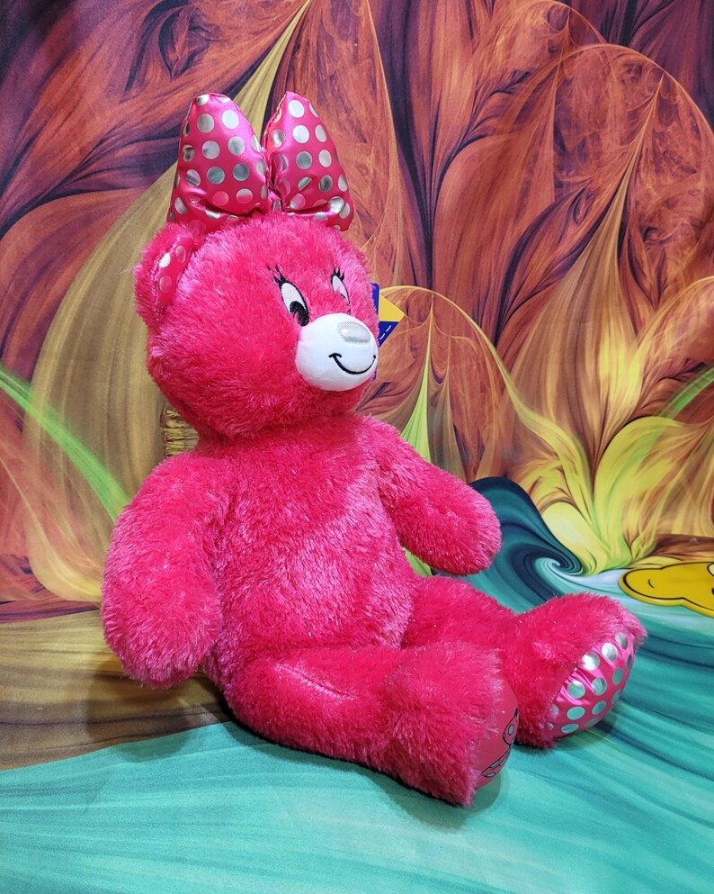 Puede incluir: Un oso de peluche rosa brillante con un gran lazo con lunares blancos. El oso tiene un hocico blanco, una cara sonriente y lunares azul claro en los pies. Est&aacute; sentado sobre una superficie verde y turquesa.