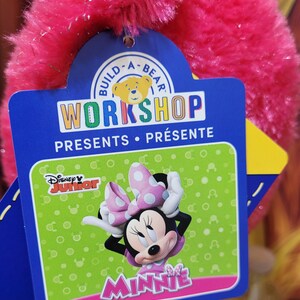 Puede incluir: Una etiqueta de Build-A-Bear Workshop con Minnie Mouse de Disney Junior. La etiqueta es azul y verde con las palabras "WORKSHOP PRESENTS" y "MINNIE" en rosa. El peluche es de color rosa vibrante.