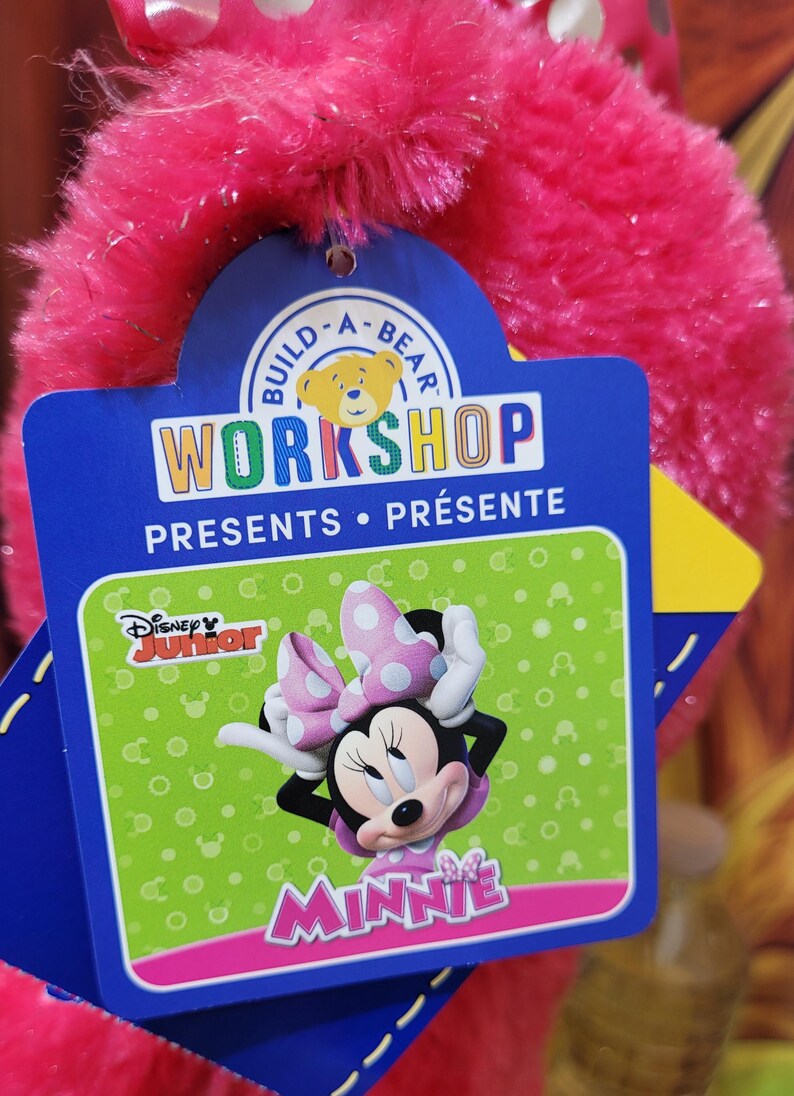 Puede incluir: Una etiqueta de Build-A-Bear Workshop con Minnie Mouse de Disney Junior. La etiqueta es azul y verde con las palabras "WORKSHOP PRESENTS" y "MINNIE" en rosa. El peluche es de color rosa vibrante.