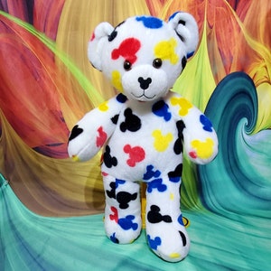 Pu&ograve; includere: Un orsacchiotto di peluche bianco con macchie colorate a forma di Mickey Mouse in rosso, giallo, blu e nero. L'orso ha un naso nero e occhi marroni. Lo sfondo presenta un design astratto colorato.