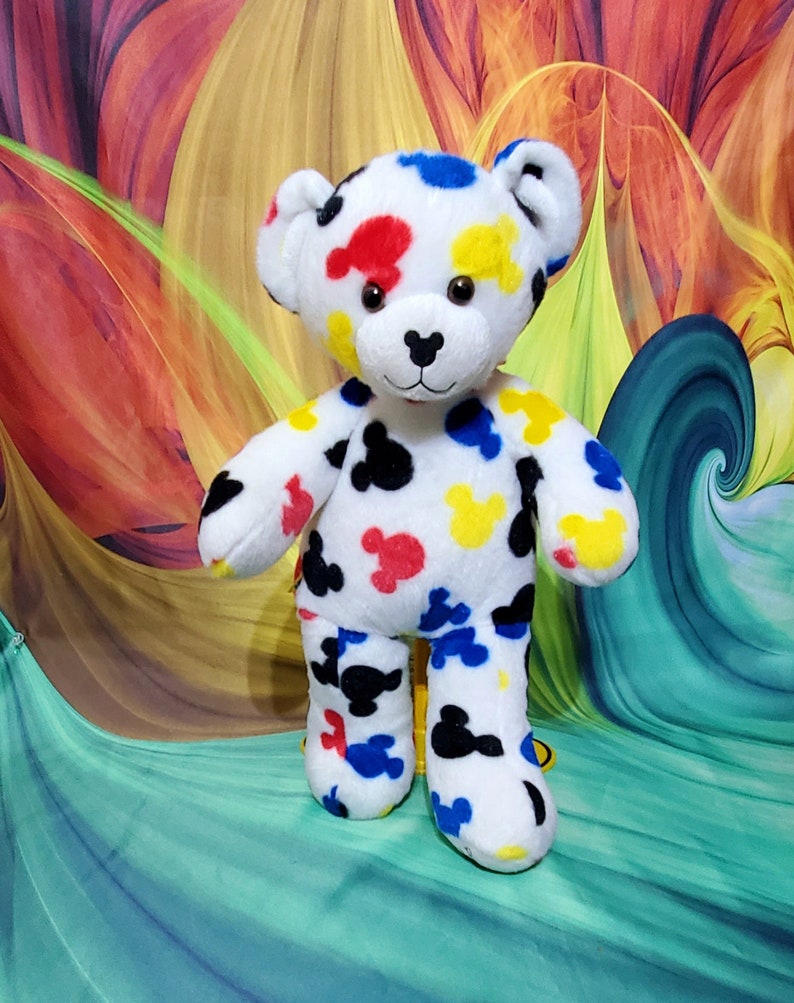 Pu&ograve; includere: Un orsacchiotto di peluche bianco con macchie colorate a forma di Mickey Mouse in rosso, giallo, blu e nero. L'orso ha un naso nero e occhi marroni. Lo sfondo presenta un design astratto colorato.