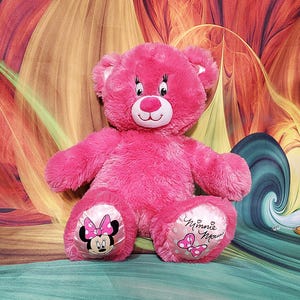 K&ouml;nnte beinhalten: Ein leuchtend rosa Teddyb&auml;r mit einem l&auml;chelnden Gesicht, wei&szlig;en Akzenten und Minnie-Maus-Designs auf den F&uuml;&szlig;en. Der B&auml;r hat eine weiche, flauschige Textur. Er sitzt auf einem bunten Hintergrund.