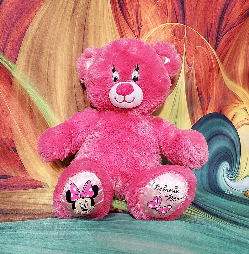 K&ouml;nnte beinhalten: Ein leuchtend rosa Teddyb&auml;r mit einem l&auml;chelnden Gesicht, wei&szlig;en Akzenten und Minnie-Maus-Designs auf den F&uuml;&szlig;en. Der B&auml;r hat eine weiche, flauschige Textur. Er sitzt auf einem bunten Hintergrund.