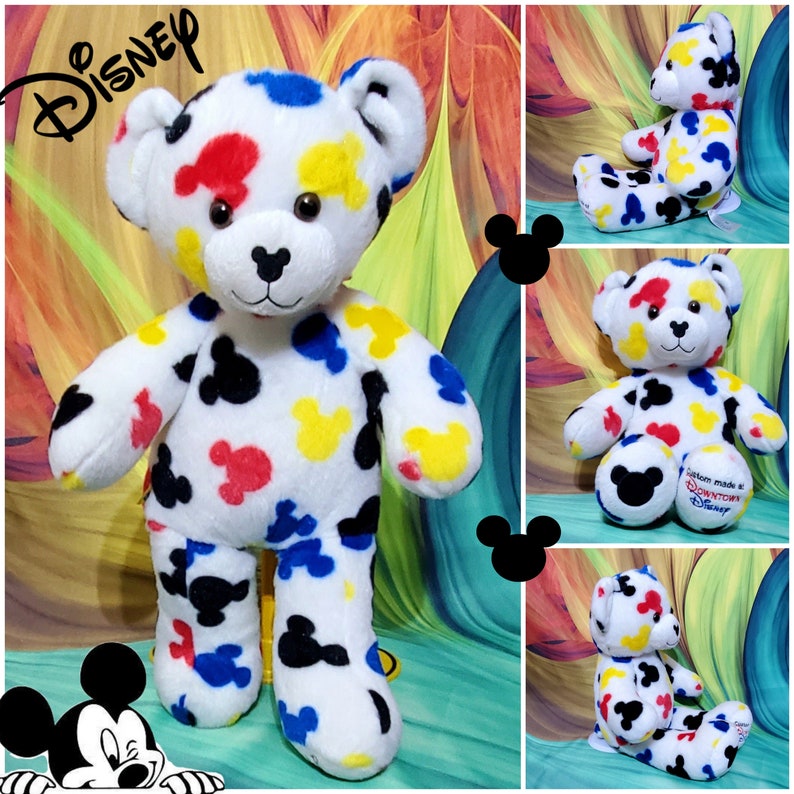 Pu&ograve; includere: Un orsacchiotto di peluche bianco con forme di testa di Mickey Mouse colorate in rosso, giallo, blu e nero. L'orso ha naso e occhi neri, con il logo Disney in alto a sinistra. Ulteriori immagini mostrano l'orso in diverse pose.