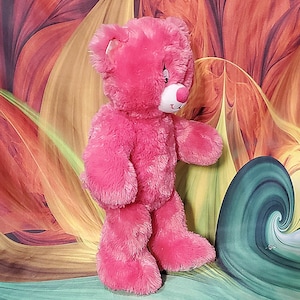 K&ouml;nnte beinhalten: Ein rosa Teddyb&auml;r mit wei&szlig;er Schnauze und rosa Nase. Der B&auml;r hat einen freundlichen Ausdruck und steht aufrecht. Der Hintergrund zeigt ein farbenfrohes abstraktes Design.