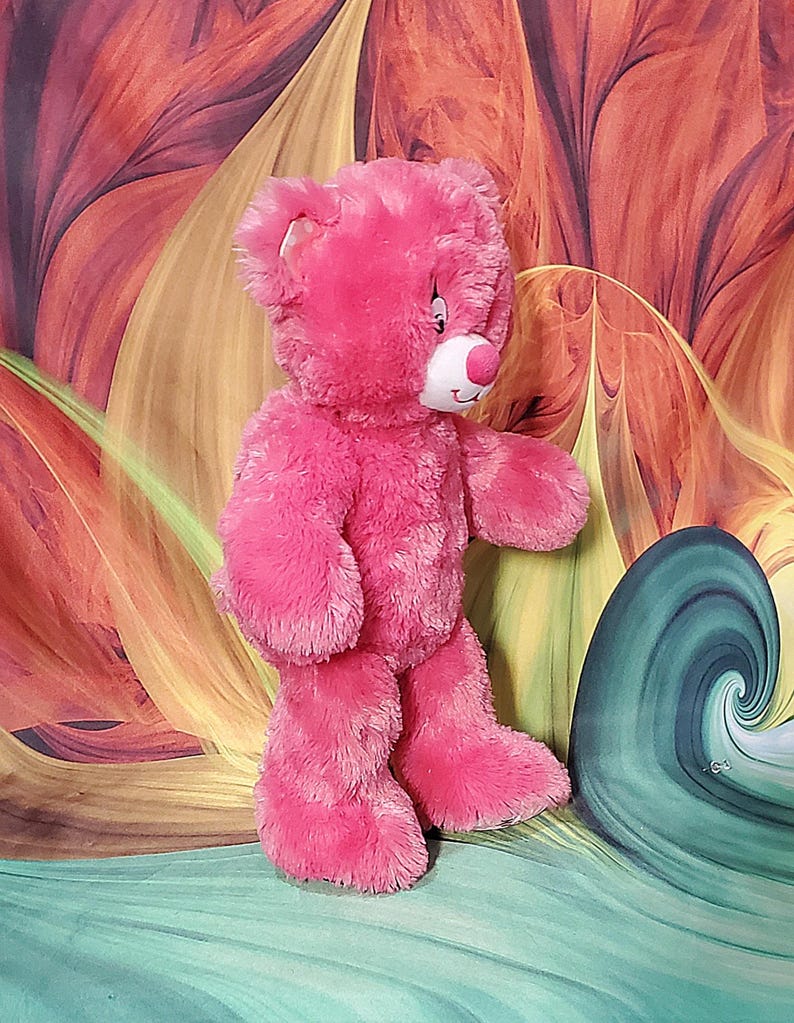 K&ouml;nnte beinhalten: Ein rosa Teddyb&auml;r mit wei&szlig;er Schnauze und rosa Nase. Der B&auml;r hat einen freundlichen Ausdruck und steht aufrecht. Der Hintergrund zeigt ein farbenfrohes abstraktes Design.