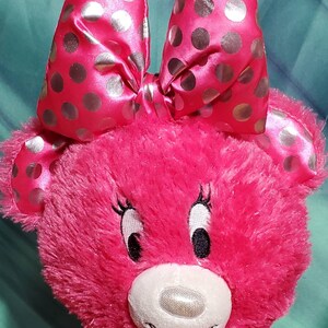 May include: A plush pink teddy bear with a large bow featuring silver polka dots. The bear has embroidered eyes, eyelashes, and a white muzzle. The bear's fur is a vibrant pink color.