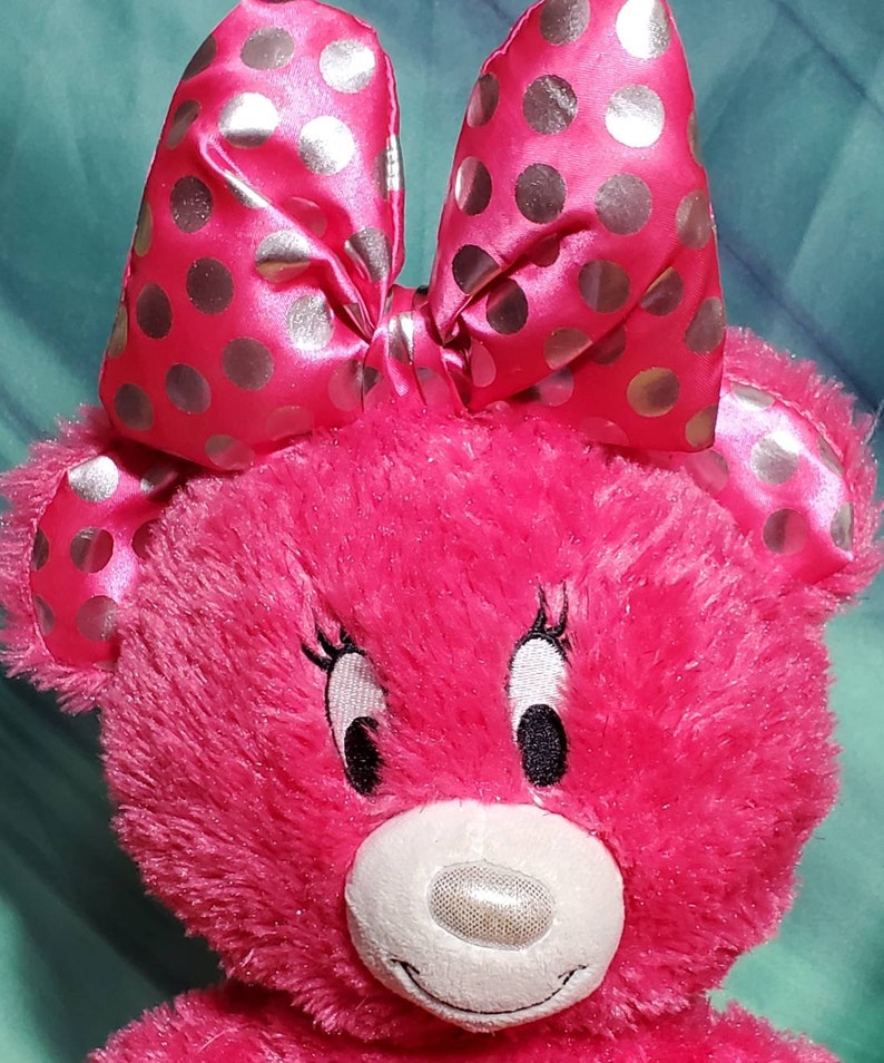 May include: A plush pink teddy bear with a large bow featuring silver polka dots. The bear has embroidered eyes, eyelashes, and a white muzzle. The bear's fur is a vibrant pink color.