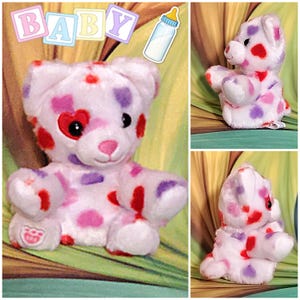May include: A white plush teddy bear with red, purple, and pink polka dots. The bear has a heart-shaped eye and a pink nose. The word "BABY" is displayed above the bear in pastel colors. A baby bottle illustration is also present.