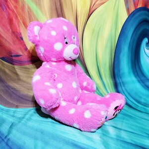May include: Pink polka dot teddy bear with white polka dots. The bear has a white face with black eyes and a pink nose. The bear is sitting on a blue and green background.