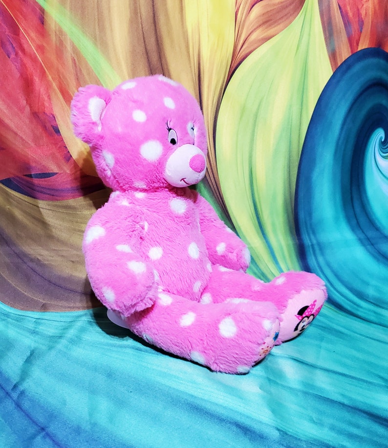 May include: Pink polka dot teddy bear with white polka dots. The bear has a white face with black eyes and a pink nose. The bear is sitting on a blue and green background.