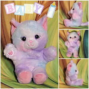 May include: A plush, pastel-colored cat toy with large black eyes, pink accents, and a paw print on one paw. The word "BABY" is displayed above the toy in colorful letter blocks, with a baby bottle illustration. The toy is designed for babies.