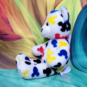 Pu&ograve; includere: Un orsacchiotto di peluche bianco con macchie colorate a forma di Mickey Mouse in rosso, giallo, blu e nero. L'orso ha un colletto rosso e un'etichetta attaccata. Lo sfondo presenta un design astratto colorato.