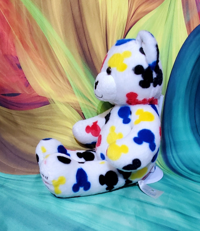 Pu&ograve; includere: Un orsacchiotto di peluche bianco con macchie colorate a forma di Mickey Mouse in rosso, giallo, blu e nero. L'orso ha un colletto rosso e un'etichetta attaccata. Lo sfondo presenta un design astratto colorato.