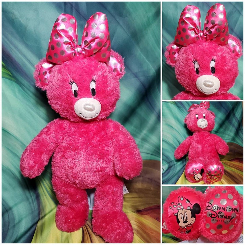 May include: A bright pink teddy bear with a Minnie Mouse theme. The bear has a large bow with silver polka dots, embroidered eyelashes, and a white muzzle. The bear's feet feature a Minnie Mouse graphic. The image also includes two round pillows with Minnie Mouse and Downtown Disney District logos.