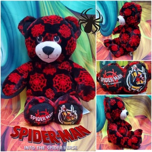 May include: A plush teddy bear featuring a black and red spiderweb pattern across its fur. The bear has a white muzzle and black nose. The soles of the feet display the text Spider-Man Into the Spider-Verse and a graphic of the character.