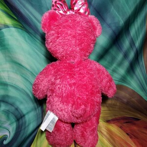 May include: A bright pink plush teddy bear with a matching bow featuring black polka dots. The bear has a soft, fuzzy texture and is viewed from the back. A white tag is attached to the bear's leg.