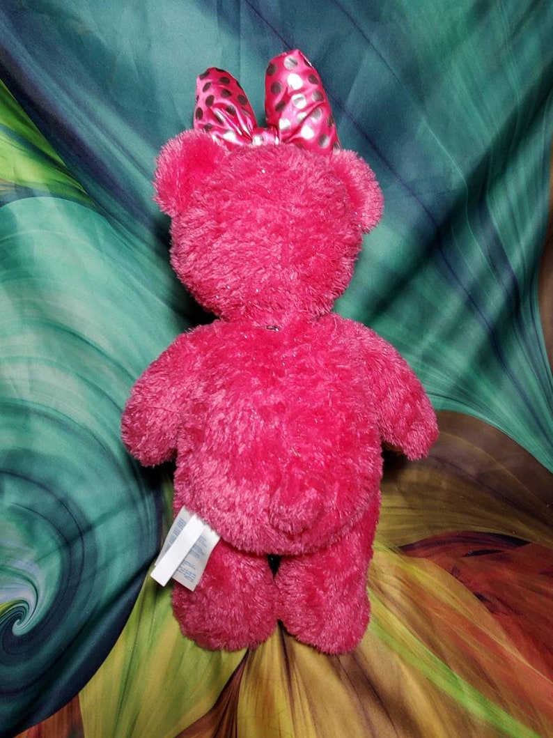 May include: A bright pink plush teddy bear with a matching bow featuring black polka dots. The bear has a soft, fuzzy texture and is viewed from the back. A white tag is attached to the bear's leg.