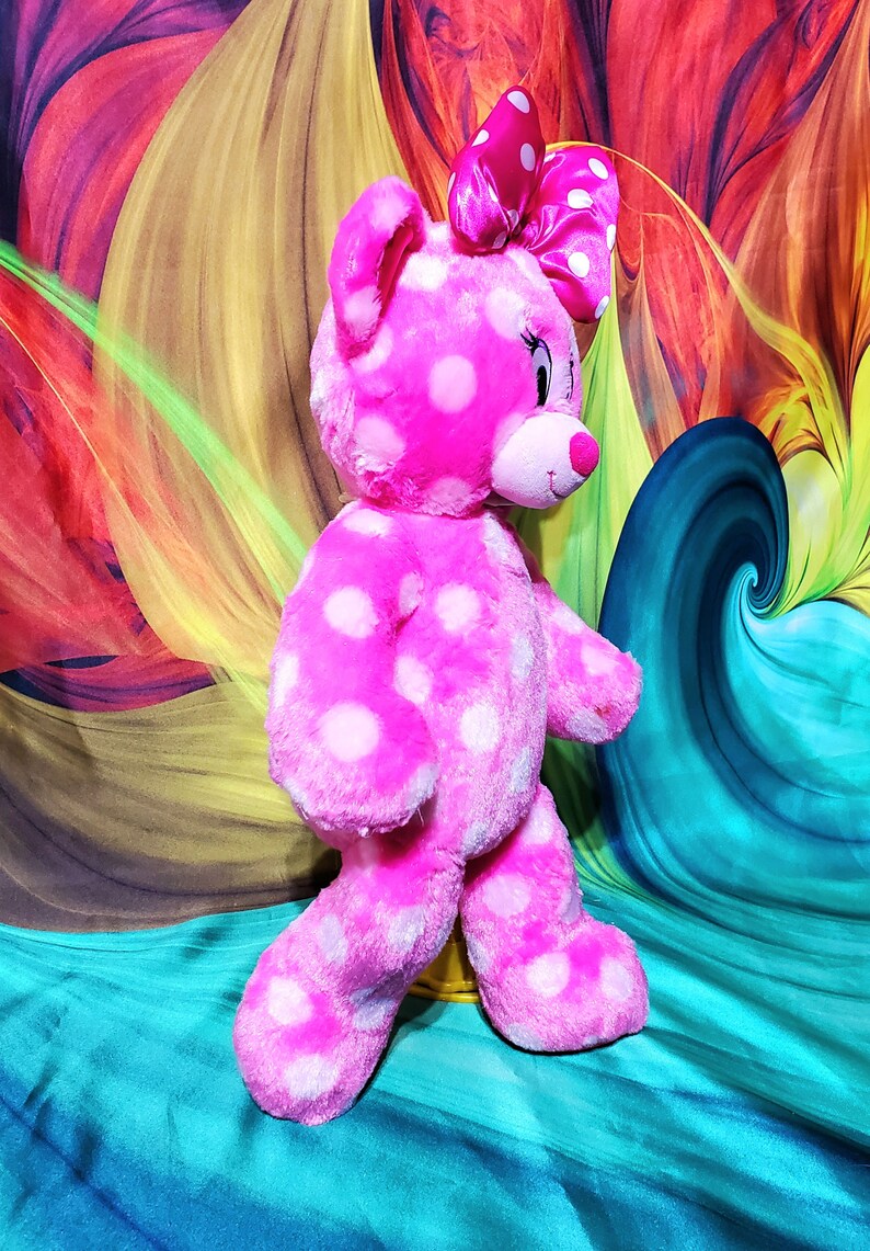 Op de afbeelding: Een roze pluche beer met witte stippen, met een bijpassende strik. De beer heeft een roze neus en staat tegen een kleurrijke, abstracte achtergrond. Dit knuffel is een schattig en knuffelbaar item.
