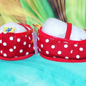 K&ouml;nnte beinhalten: Ein Paar rote Babyschuhe mit wei&szlig;en Punkten. Die Schuhe haben einen roten Riemen und eine Schleife. Die Schuhe sind neu und haben ein Etikett mit einem Pfotenabdruck. Auf farbenfrohem Hintergrund.