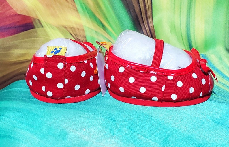 K&ouml;nnte beinhalten: Ein Paar rote Babyschuhe mit wei&szlig;en Punkten. Die Schuhe haben einen roten Riemen und eine Schleife. Die Schuhe sind neu und haben ein Etikett mit einem Pfotenabdruck. Auf farbenfrohem Hintergrund.