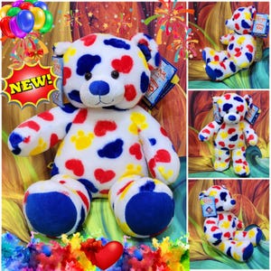 May include: A white plush teddy bear with red hearts, yellow paw prints, and blue spots. The bear has blue accents on its nose, ears, and feet. A "NEW!" sticker is visible. The bear is a stuffed animal.