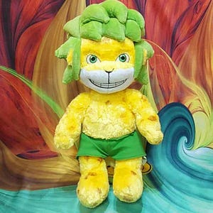 May include: A plush toy lion with a yellow body, green hair, and green shorts. The lion has a smiling face with green eyes. The background is a colorful abstract design.
