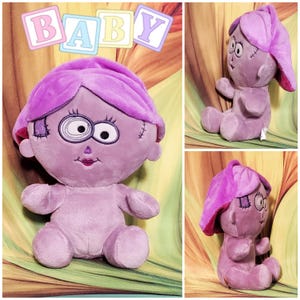 May include: A plush doll in shades of purple, featuring a stylized face with large eyes and stitched details. The doll has a distinctive head covering and is designed to resemble a baby. The word "BABY" is displayed above the doll.