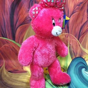 Puede incluir: Un oso de peluche rosa brillante con un lazo a juego con lunares plateados. El oso tiene un hocico blanco, ojos negros y una sonrisa cosida. Est&aacute; de pie sobre un fondo colorido y abstracto.