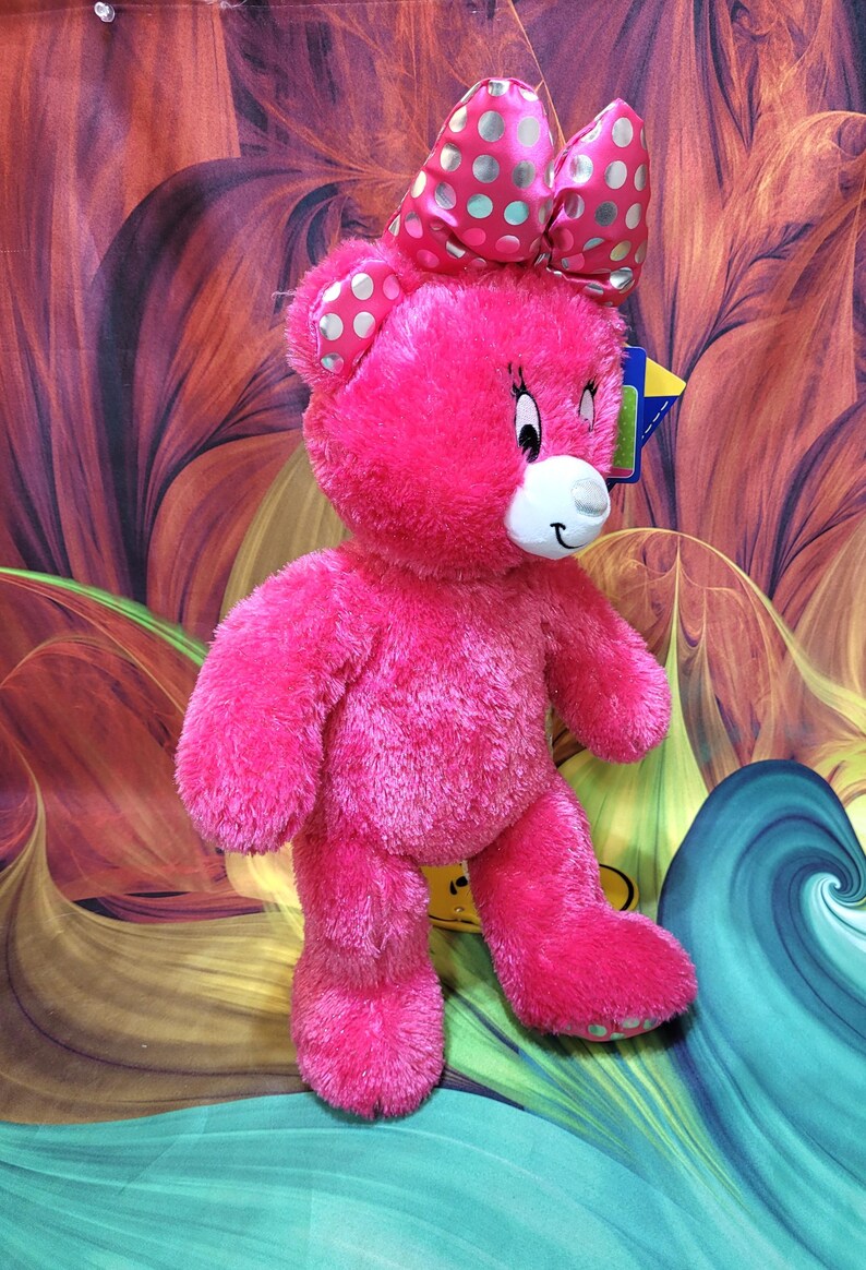 Puede incluir: Un oso de peluche rosa brillante con un lazo a juego con lunares plateados. El oso tiene un hocico blanco, ojos negros y una sonrisa cosida. Est&aacute; de pie sobre un fondo colorido y abstracto.
