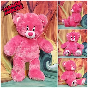 K&ouml;nnte beinhalten: Ein pinkfarbener Minnie Mouse Teddyb&auml;r mit wei&szlig;en Akzenten an den Ohren und einer roten Nase. Der B&auml;r hat gestickte Augen und einen l&auml;chelnden Mund. Auf den F&uuml;&szlig;en des B&auml;ren sind Minnie Mouses Bild und die Worte "Minnie Mouse" abgebildet. Ein Stofftier.