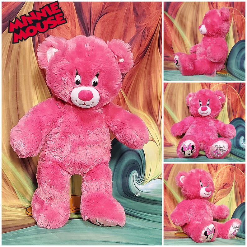 K&ouml;nnte beinhalten: Ein pinkfarbener Minnie Mouse Teddyb&auml;r mit wei&szlig;en Akzenten an den Ohren und einer roten Nase. Der B&auml;r hat gestickte Augen und einen l&auml;chelnden Mund. Auf den F&uuml;&szlig;en des B&auml;ren sind Minnie Mouses Bild und die Worte "Minnie Mouse" abgebildet. Ein Stofftier.