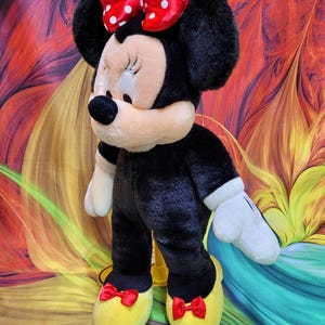 K&ouml;nnte beinhalten: Eine Pl&uuml;schpuppe von Minnie Mouse, mit einem schwarzen K&ouml;rper, wei&szlig;en Handschuhen und gelben Schuhen mit roten Schleifen. Ihr Gesicht ist hellbraun mit einer schwarzen Nase und geschlossenen Augen. Eine rote Schleife mit wei&szlig;en Punkten schm&uuml;ckt ihren Kopf. Die Puppe ist ca. 46 cm gro&szlig;.