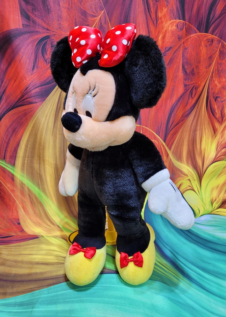 K&ouml;nnte beinhalten: Eine Pl&uuml;schpuppe von Minnie Mouse, mit einem schwarzen K&ouml;rper, wei&szlig;en Handschuhen und gelben Schuhen mit roten Schleifen. Ihr Gesicht ist hellbraun mit einer schwarzen Nase und geschlossenen Augen. Eine rote Schleife mit wei&szlig;en Punkten schm&uuml;ckt ihren Kopf. Die Puppe ist ca. 46 cm gro&szlig;.