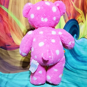 May include: Pink plush teddy bear with white polka dots. The bear is standing on a colorful background.