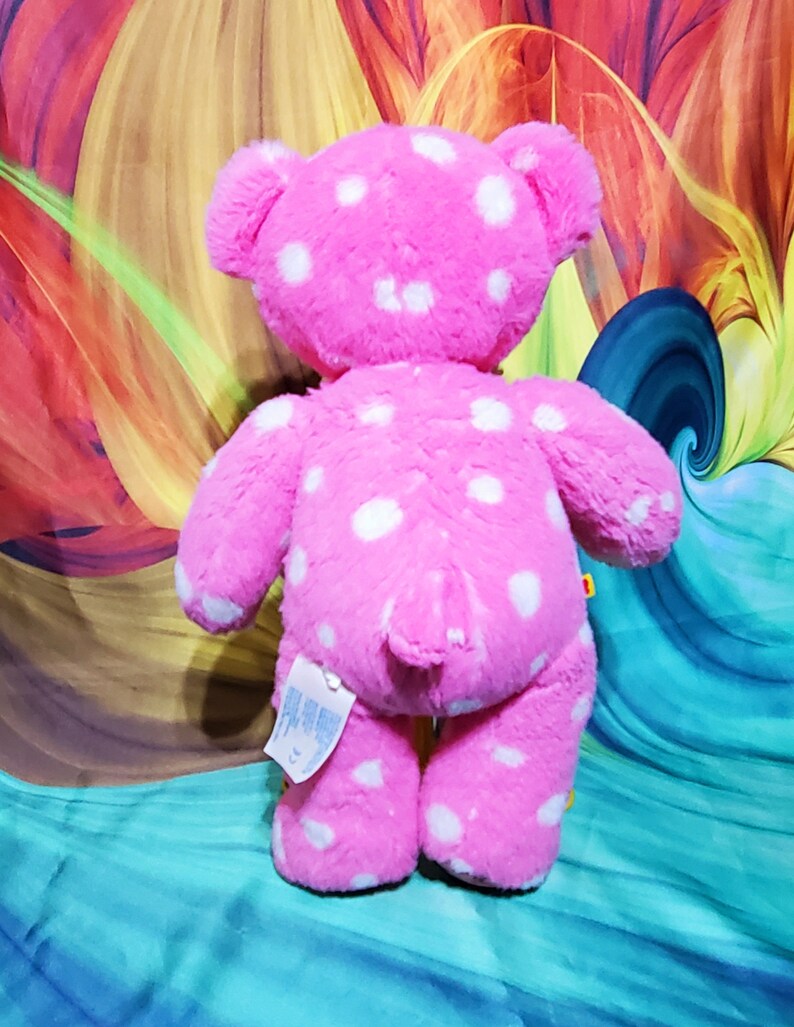 May include: Pink plush teddy bear with white polka dots. The bear is standing on a colorful background.