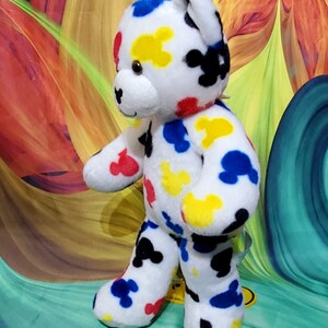 Pu&ograve; includere: Un orsacchiotto di peluche bianco con macchie colorate a forma di Mickey Mouse rosse, gialle, blu e nere. L'orso ha muso, orecchie e zampe bianche. Lo sfondo &egrave; un design astratto colorato.