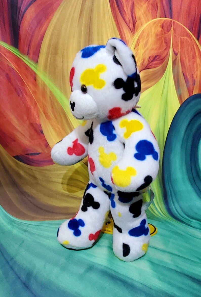 Pu&ograve; includere: Un orsacchiotto di peluche bianco con macchie colorate a forma di Mickey Mouse rosse, gialle, blu e nere. L'orso ha muso, orecchie e zampe bianche. Lo sfondo &egrave; un design astratto colorato.